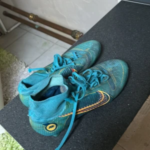 Blå Nike fotbollsskor med snörning - Säljer ett par blå Nike fotbollsskor med gula och röda detaljer samt snörning. Skorna har en rund tå och är tillverkade i syntetmaterial. Perfekta för dig som vill sticka ut på planen!