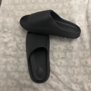 Svarta slides sandaler - Säljer ett par svarta slides med öppen tå och platt sula. De har en minimalistisk design och är tillverkade i syntetmaterial. Perfekta för sommaren eller att ha hemma.