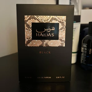 Hawas Black Eau de Parfum för honom - En elegant parfymflaska i svart glas med silverdetaljer och text på både engelska och arabiska. Doften är Eau de Parfum och flaskan rymmer 100 ml. Förpackningen har ett mönster i ormskinnslook och svart, vilket ger en lyxig känsla. Kvar i 97ml 