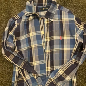 Ralph Lauren flanell skjorta - Säljer en klassisk blårutig skjorta från Ralph Lauren med lång ärm och knappar framtill. Skjortan har en broderad rosa logga på bröstet och är tillverkad i mjuk bomull. Perfekt för dig som gillar stilrena och tidlösa plagg.
