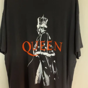 Svart Queen t-shirt med tryck - Cool svart t-shirt från Queen Official Merch med stort tryck av Freddie Mercury i krona och mantel samt texten 'QUEEN' i rött framtill. Tillverkad i 100% bomull och har en klassisk passform med korta ärmar. Perfekt för fans av bandet! Aldrig haft på mig den själv.