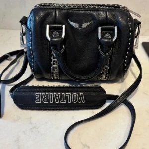 Svart axelväska från Zadig & Voltaire - Cools svart axelväska i skinn från Zadig & Voltaire med silverfärgade nitar och detaljer. Väskan har dragkedja upptill, dubbla handtag och justerbar axelrem med logga. Perfekt för att lyfta din outfit med en edgy touch. Den har rostat lite men syns knappast (se bild)