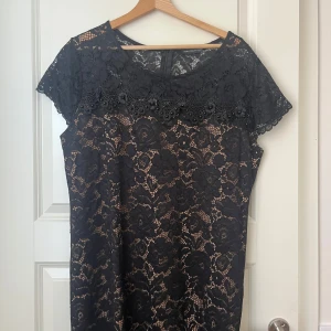 Svart spetsklänning med blommönster - Svart klänning med vackert blommigt spetsmönster och dekorativa pärlor vid halsringningen. Klänningen har korta ärmar och är fodrad i beige för en snygg kontrast. Perfekt för dig som gillar eleganta detaljer och klassisk stil.