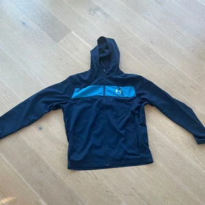 Svart träningsjacka med blå detalj från Under Armour - Snygg svart träningsjacka från Under Armour med blå panel över bröstet och vit logga. Jackan har huva, dragkedja och två sidofickor. Perfekt för träning eller sportiga dagar.
