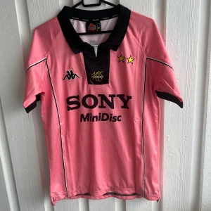 Juventus retro fotbollströja Zidane #21 - Säljer en rosa Juventus retro fotbollströja med svart krage och detaljer, tryckta stjärnor och sponsorlogga på bröstet. På ryggen står det Zidane och nummer 21. Perfekt för samlare eller fans!