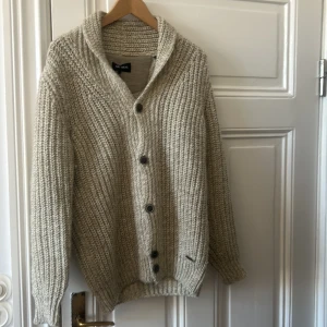 Beige stickad kofta med knappar - Säljer en chunky beige stickad kofta. Koftan har grovstickad struktur, stora knappar framtill och en bred krage. Perfekt för lager-på-lager och håller dig varm under kyliga dagar. Mcneal