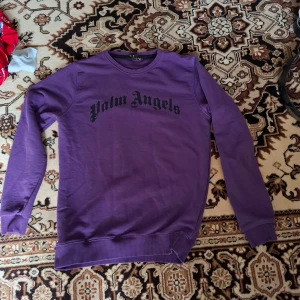 Lila sweatshirt från Palm Angels - Säljer en lila sweatshirt från Palm Angels med svart tryck över bröstet. Tröjan har rund halsringning, långa ärmar och ribbade muddar vid ärmslut och nederkant. Perfekt för en avslappnad och trendig stil.