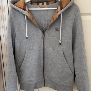 Grå hoodie med dragkedja från Burberry - Säljer en grå hoodie från Burberry med dragkedja i fint skick! Perfekt nu inför sommaren som matchar bra med linnebyxor. 100% ÄKTA