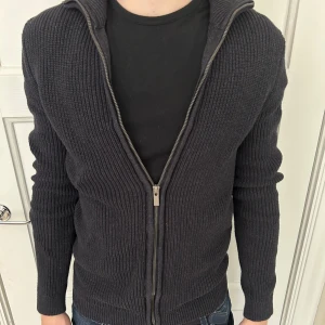 Mörkgrå stickad kofta med dragkedja från Dressmann - Säljer en mörkgrå stickad kofta från Dressmann i storlek S. Tröjan har hel dragkedja framtill, ribbad struktur och hög krage. 