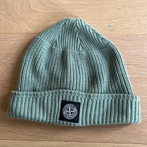 Grön lyxig ribbstickad mössa från Stone Island - Snygg grön ribbstickad mössa från Stone Island  med klassisk svart logotyp framtill. Perfekt accessoar för kyligare dagar och enkel att matcha med olika outfits. Mössan är ganska tunn men snygg för en lyxig look 