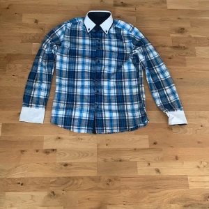 Blårutig skjorta med vita detaljer - Snygg blårutig skjorta med vita manschetter och krage. Skjortan har lång ärm, knappar framtill och klassisk passform. Perfekt för dig som gillar en stilren look med lite edge.