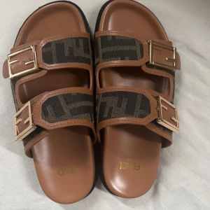 Bruna sandaler från Fendi med monogram - Snygga sandaler från Fendi med två breda remmar i brunt och Fendi-monogram. Guldiga spännen och bekväm platt sula. Perfekta för sommaren och ger en lyxig touch till din outfit. Aldrig använda utomhus 