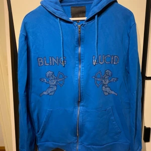 Blå hoodie med dragkedja från Blinglucid - Säljer en blå hoodie från Blinglucid i storlek L. Tröjan har dragkedja, huva och fickor framtill. På bröstet finns glittriga tryck med texten 'BLING LUCID' och två amor-motiv. Perfekt för en avslappnad stil.