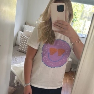 Vit t-shirt från Stella McCartney Kids med tryck - Vit t-shirt från Stella McCartney Kids med stort lila tryck framtill och texten 'SHELL McCARTNEY'. Aldrig använd 