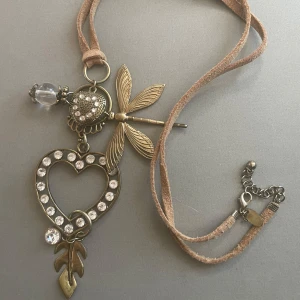 Halsband med hjärta, trollslända och strass - Halsband med antikguldfärgad kedja, stort hjärthänge dekorerat med klara strasstenar, trollslända och lövdetalj. Hängsmycket har även en glaskula och dekorativa mönster. Med läderrem (fakeläder)