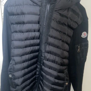 Moncler cardigan - Säljer en svart pufferjacka från Moncler med quiltade partier framtill och på huvan. Jackan har dragkedja, ficka med dragkedja på ärmen och Moncler-logga på axeln. Insidan har en serietidningsdetalj och färgade dragkedjor.