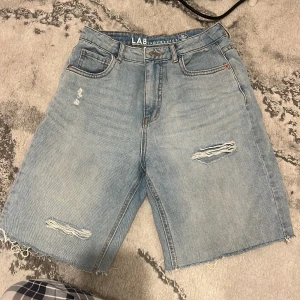 Ljusblå jeansshorts från LAB Industries - Ljusblå jeansshorts med råa kanter och slitna detaljer framtill. Klassisk femficksmodell med knapp och dragkedja. Perfekta för en avslappnad stil.