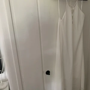 Vit långklänning med tunna axelband - Säljer en elegant vit långklänning med tunna axelband och v-ringning. Klänningen har en enkel och stilren design med lätt utställd passform och knytning i ryggen. Perfekt för sommarens alla tillfällen.