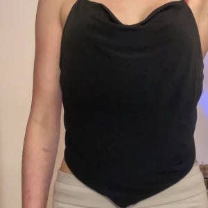 Svart croppad linnetopp med tunna axelband - Säljer en svart croppad linnetopp med tunna axelband som korsas i ryggen. Toppen har en snygg draperad halsringning och spetsig nederkant framtill. Perfekt för att styla med jeans eller kjol.