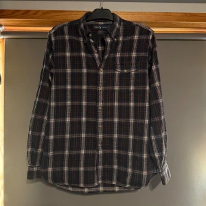 Rutig skjorta från Ralph Lauren - Snygg rutig skjorta från Ralph Lauren i mörka färger med inslag av vitt och rött. Klassisk krage, knappstängning och en bröstficka. Perfekt för dig som gillar en tidlös och stilren look.