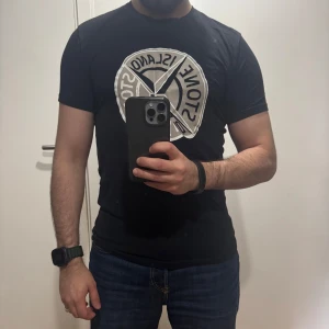 Svart t-shirt från Stone Island med stor logga - Säljer en svart t-shirt från Stone Island med en stor, rund logga i vitt och grått på bröstet. T-shirten har klassisk passform och korta ärmar. Perfekt för dig som gillar streetwear och vill sticka ut med en ikonisk design.