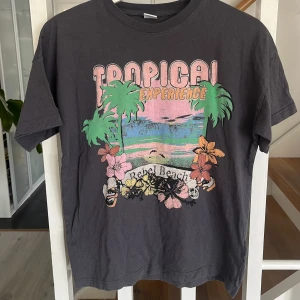 Grå t-shirt med tropiskt tryck från Lindex - Säljer en grå t-shirt från Lindex med färgglatt tryck av palmer, blommor och texten 'Tropical Experience Rebel Beach' på framsidan. T-shirten har korta ärmar och är i mjuk bomull. Perfekt för sommardagar och strandhäng!