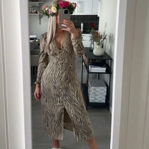 Adoore klänning  - Säljer en långklänning från Adoore med zebramönster i beige och brunt. Klänningen har lång ärm, v-ringning och knappar längs hela framsidan. Perfekt för sommarens tillfällen och midsommarfirande. Den är använd ett fåtal gånger och ser ut och vara ny💕