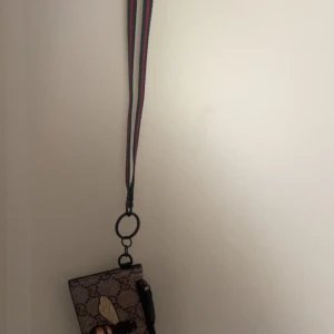 Gucci korthållare med nyckelband - Säljer en snygg korthållare från Gucci med klassiskt GG-mönster och bi-detalj på framsidan. Korthållaren har flera fack och ett svart nyckelband med röd-gröna ränder. Perfekt för kort och nycklar.