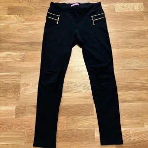 Svarta leggings med guldfärgade dragkedjor från Sofia Paris - Svarta leggings från Sofia Paris med två dekorativa guldfärgade dragkedjor framtill. Byxorna har en tight passform och är tillverkade i ett stretchigt material som sitter skönt på kroppen. Perfekta för en stilren och enkel look.