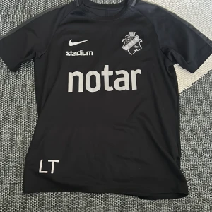 Svart AIK fotbollströja från Nike - Säljer en svart AIK fotbollströja från Nike med klubbmärke, sponsortryck och Dri-FIT-material. Tröjan har korta ärmar och tryck från Notar, Stadium och SVEA Ekonomi. Perfekt för dig som vill visa ditt AIK-stöd på träning eller match.