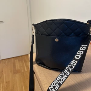 Svart quiltad axelväska med textad rem - Snygg svart axelväska med quiltad ovandel och bred axelrem med vit text. Väskan har guldfärgade metalldetaljer och en knapp framtill. Perfekt för dig som vill ha en trendig accessoar med plats för det viktigaste.