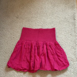 Rosa volangkjol från Lindex - Supersöt rosa kjol från Lindex med bred smock i midjan och volangig, puffig kjoldel. Perfekt för dig som gillar färg och vill ha en bekväm men ändå snygg kjol. Kjolen är kort och har en luftig känsla.