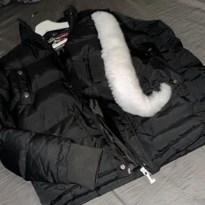 Svart dunjacka från Moose Knuckles - Säljer en svart dunjacka från Moose Knuckles med fluffig vit fuskpäls på insidan av luvan. Jackan har dragkedja, tryckknappar och ribbade muddar vid ärmsluten. Perfekt för kalla dagar och riktigt snygg streetstil. Betala genom Swish 