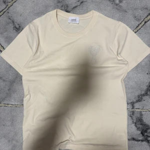 Beige t-shirt från Ami Paris med hjärtdetalj - Snygg beige t-shirt från Ami Paris med ett broderat hjärta och bokstaven A på bröstet. Klassisk rund halsringning och korta ärmar. Perfekt för en clean och stilren look.