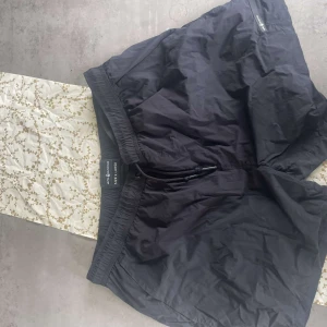 Svarta träningsshorts från Sail Racing - Svarta shorts från Sail Racing med elastisk midja och dragsko. Shortsen har fickor med dragkedja och är tillverkade i ett lätt syntetmaterial, perfekta för träning eller aktiva dagar.