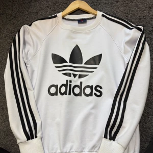 Vit Adidas sweatshirt med svarta ränder - Säljer en vit Adidas sweatshirt med klassiska svarta ränder längs ärmarna och stor logga framtill. Tröjan har rund halsringning och långa ärmar med muddar. Perfekt för en sportig och avslappnad stil.