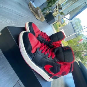 Nike Air Jordan 1 mid - Säljer ett par klassiska Nike Air Jordan 1 mid i svart och rött med vita sulor. Skorna har högt skaft, röd yttersula och ikonisk Jordan-logga på sidan. Perfekta för dig som gillar streetwear och sneakerskultur.