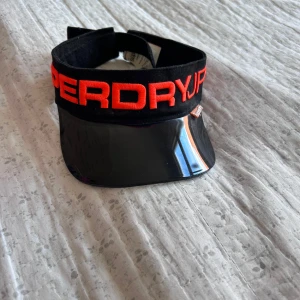 Svart visirkeps från Superdry - Snygg svart visirkeps från Superdry med bredt svart skärm och tydlig röd broderad logga framtill. Justerbar passform med kardborreband. Perfekt för soliga dagar och sportiga outfits.