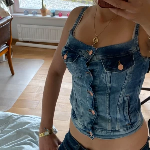Jeans korsett-topp med knappar - Säljer en snygg jeans korsett-topp med smala axelband och dekorativa fickor framtill. Toppen har knappar hela vägen ner och en figurnära passform. Perfekt för dig som vill ha en cool och trendig look. Jag har sytt om den lite själv förhand. 