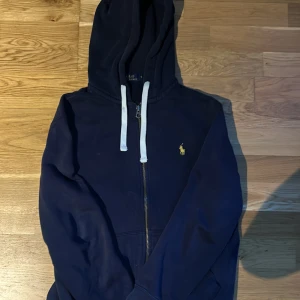 Mörkblå hoodie från Polo Ralph Lauren - Säljer en mörkblå hoodie från Polo Ralph Lauren med dragkedja och vit snörning i huvan. Klassisk gul logga broderad på bröstet. Perfekt för en avslappnad stil och enkel att matcha med det mesta.