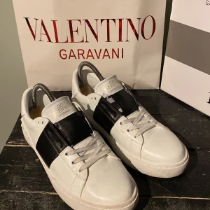 Valentino Garavani vita och svarta sneakers - Säljer ett par snygga vita sneakers från Valentino Garavani med bred svart detalj över sidan. Skorna har rund tå, platt sula och klassisk snörning. Perfekta för dig som gillar stilrena och exklusiva sneakers.
