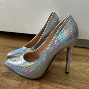 Holografiska pumps med ormskinnsmönster - Säljer ett par holografiska pumps med ormskinnsmönster och hög klack. Skorna har spetsig tå och skimrar i regnbågens färger beroende på ljuset. Perfekta för att sticka ut och ge en futuristisk touch till outfiten.  De är oanvända står storlek 35 men snarare 36