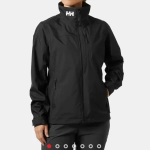 Svart vindjacka från Helly Hansen - Svart vindjacka från Helly Hansen med hög krage och dragkedja framtill. Jackan har tre fickor med dragkedja och diskreta vita loggor på bröstet, ärmen och nacken. Perfekt för blåsiga dagar och enkel att matcha.