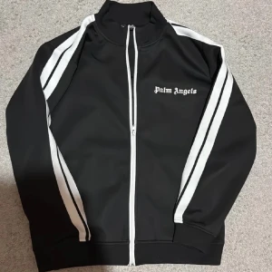 Svart track jacket från Palm Angels - Snygg svart track jacket från Palm Angels med vita ränder längs ärmarna och dragkedja framtill. Jackan har en hög krage och Palm Angels-logga på bröstet. Perfekt för en sportig look.