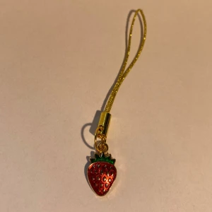 Jordgubbsberlock med guldfärgad rem - Söt accessoar med en liten jordgubbsberlock i rött och grönt, fäst på en glittrig guldfärgat hänge. Perfekt att hänga på mobilen, väskan eller nyckelknippan. Berlocken har detaljer i guld och skinande yta. Perfekt under sommaren!