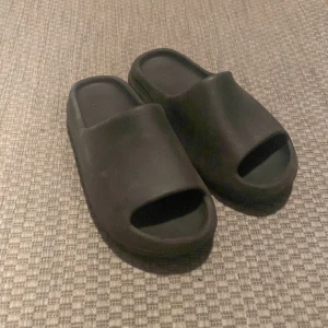 Svarta slides sandaler - Säljer ett par svarta slides sandaler med öppen tå och platt sula. De är tillverkade i syntetmaterial och har en enkel, modern design som passar perfekt för sommaren. Lite smutsiga men rengörs innan dom blir sålda 