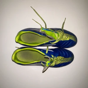 Blåa Adidas fotbollsskor med gula detaljer - Säljer ett par blåa Adidas fotbollsskor med gula snören och innersula. Skorna har vita ränder på sidorna och dobbar under sulan för bra grepp på planen. Perfekta för dig som vill sticka ut på fotbollsplanen!