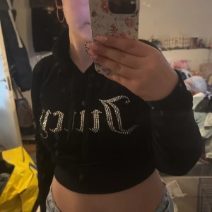 Svart croppad hoodie från Juicy Couture - Säljer en svart croppad hoodie från Juicy Couture med glittrig text framtill. Tröjan har långa ärmar, huva och dragsko. Perfekt för dig som gillar en trendig och bekväm stil.
