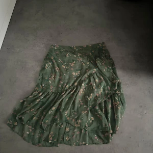 Grön blommig kjol från Vero Moda - Säljer en grön långkjol från Vero Moda med blommigt mönster. Kjolen har volangdetaljer och är tillverkad i ett lätt och luftigt material, perfekt för varmare dagar.