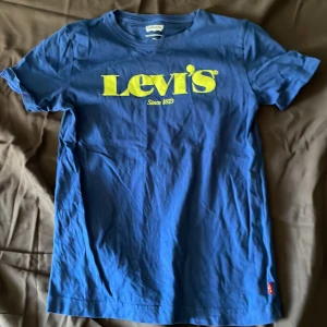 Blå t-shirt från Levi's med gul logga - Säljer en blå t-shirt från Levi's med stor gul logotyp och texten 'Since 1873' på bröstet. Klassisk rund halsringning och korta ärmar. Perfekt för dig som gillar en enkel men snygg stil.